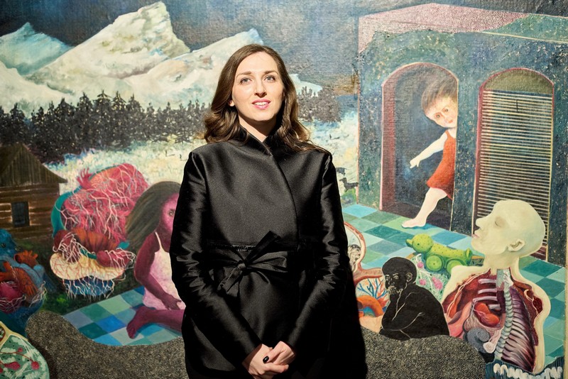 Umetnica stoji ispred velike nadrealističke slike sa figurativnim motivima, planinskim pejzažom i antropomorfnim elementima na izložbi u galerijskom prostoru.