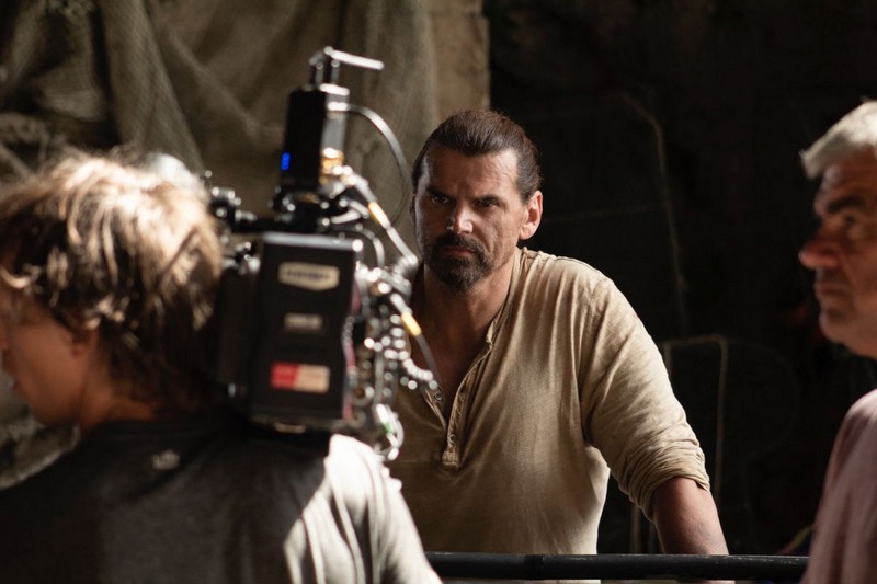 Muškarac sa vezanom kosom i bradom stoji ispred filmske kamere na setu, dok ga ekipa snima u zatvorenom prostoru sa prigušenim osvetljenjem.