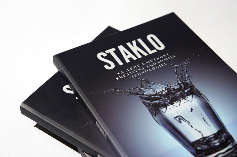 Knjiga „Staklo: nasleđe, umetnost, kreativna ekonomija, tehnologija“ sa tamnom naslovnicom na kojoj je prikazana čaša sa prskanjem vode, položena na beloj podlozi.