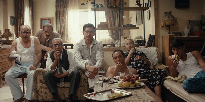 Grupa ljudi sedi u dnevnoj sobi i zajedno gleda televiziju, dok jedu voće i deluju zamišljeno, scena iz filma prikazanog na Austrijskom filmskom festivalu 2026.