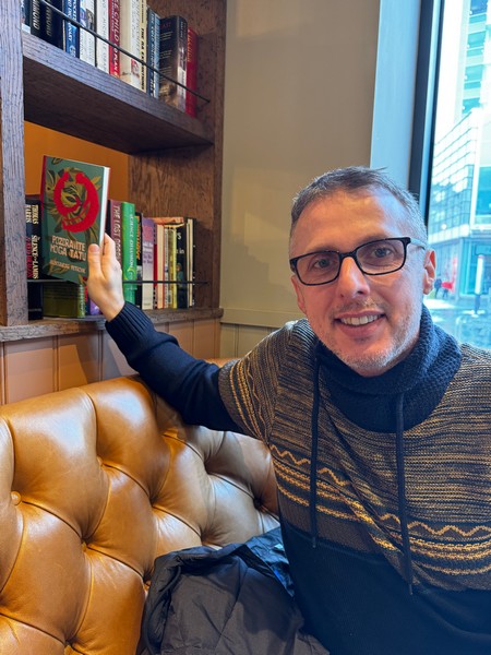 Autor drži roman „Pozdravite moga tatu“ u kafiću pored police sa knjigama, promocija savremene domaće književnosti