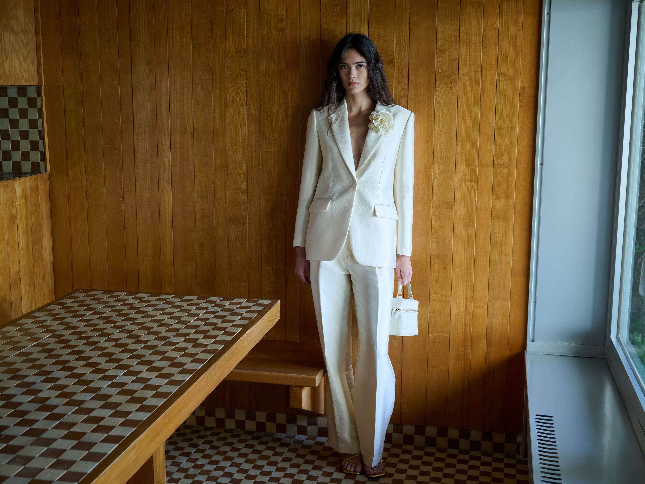 Elegantna žena u krem belom odelu sa širokim pantalonama i strukturiranim sakoom sa cvetnim detaljem, stoji u minimalističkom enterijeru sa drvenim zidovima i geometrijskim podom, držeći malu torbu.