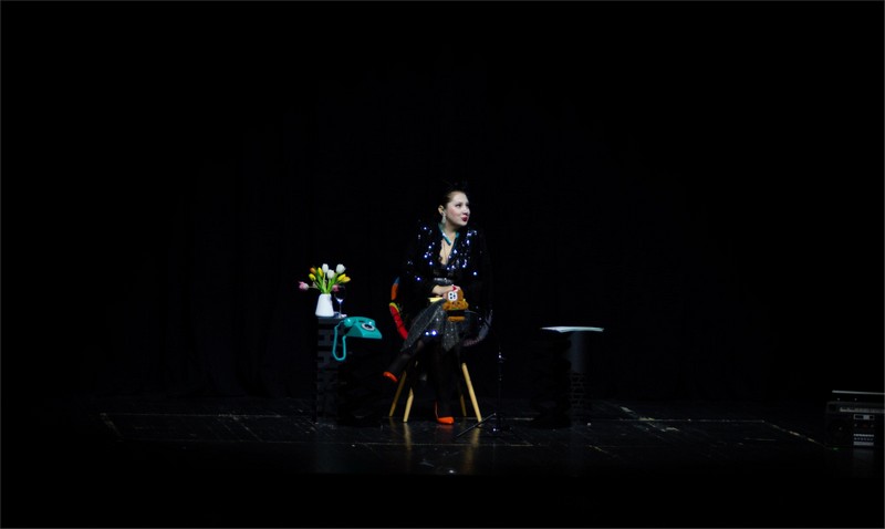 Glumica sedi na stolici na pozorišnoj sceni tokom monodrame „Seansa“, sa mikrofonom ispred sebe i minimalističkom scenografijom u mraku.