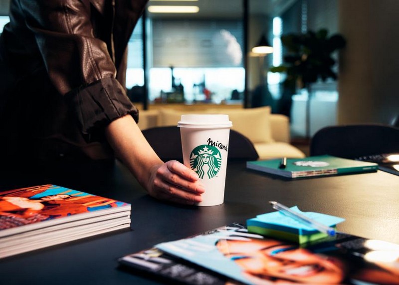 Ruka drži Starbucks čašu kafe na stolu okruženom modnim magazinima i notesima u modernom kancelarijskom okruženju.