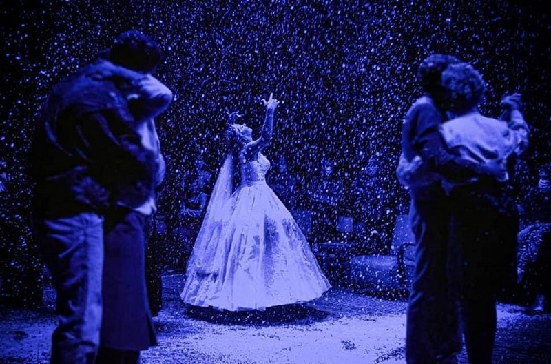 Glumci na sceni tokom izvođenja predstave „Tri zime“, u atmosferi plavog svetla i padajućeg veštačkog snega, dok centralna ženska figura u beloj haljini podiže ruke, a parovi oko nje stoje zagrljeni.