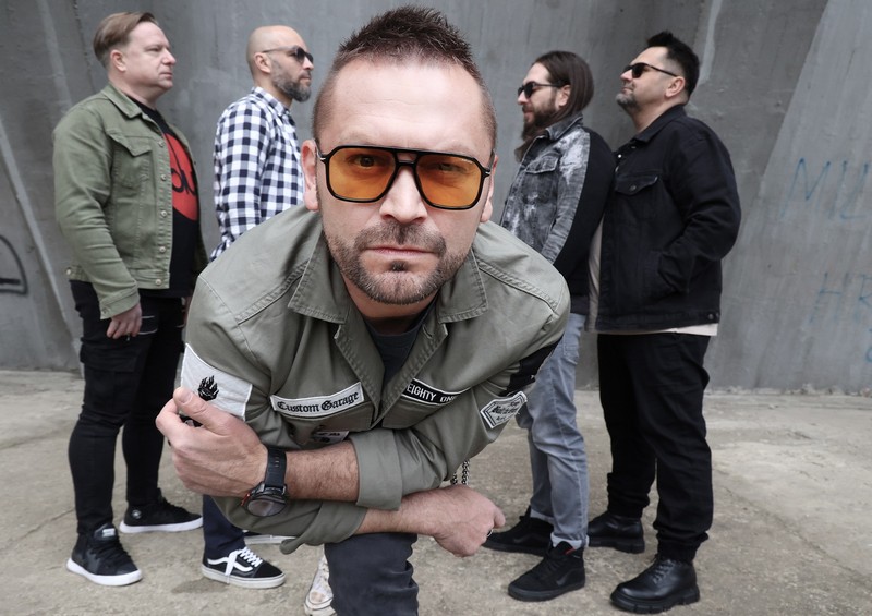 Muzički bend pozira ispred betonskog zida, dok frontmen u prvom planu, sa narandžastim naočarima i zelenom jaknom, gleda direktno u kameru, a ostali članovi stoje iza njega u profilu.