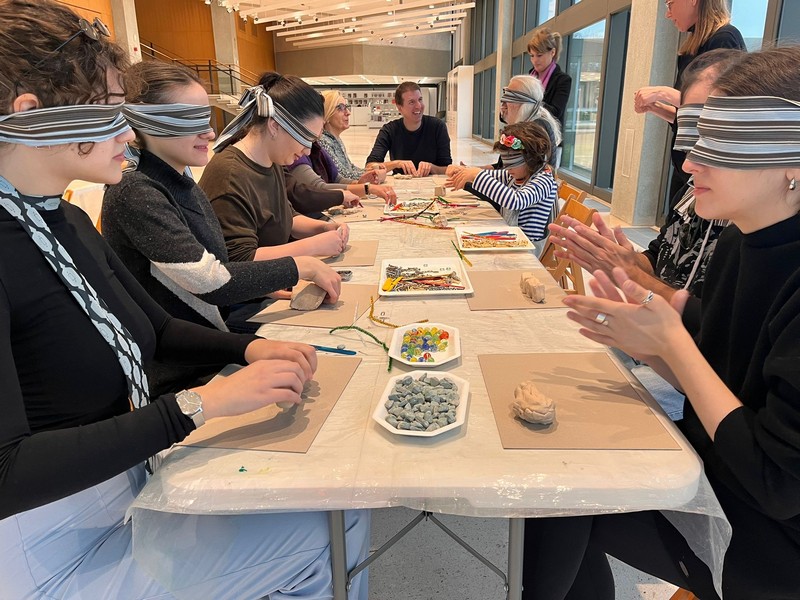 Učesnici inkluzivne radionice sede za stolom sa povezima preko očiju i kroz dodir oblikuju materijale, istražujući umetnost kroz višečulno iskustvo u muzeju.