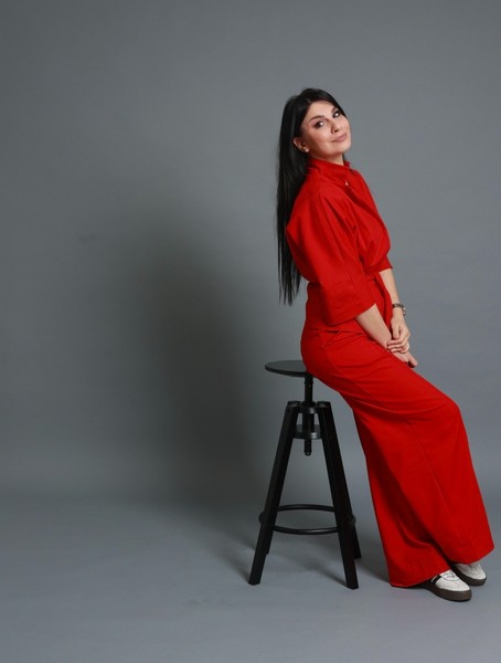 Žena sa dugom tamnom kosom u crvenom odelu sedi na barskoj stolici i blago se osmehuje u minimalističkom studijskom okruženju sa sivom pozadinom.