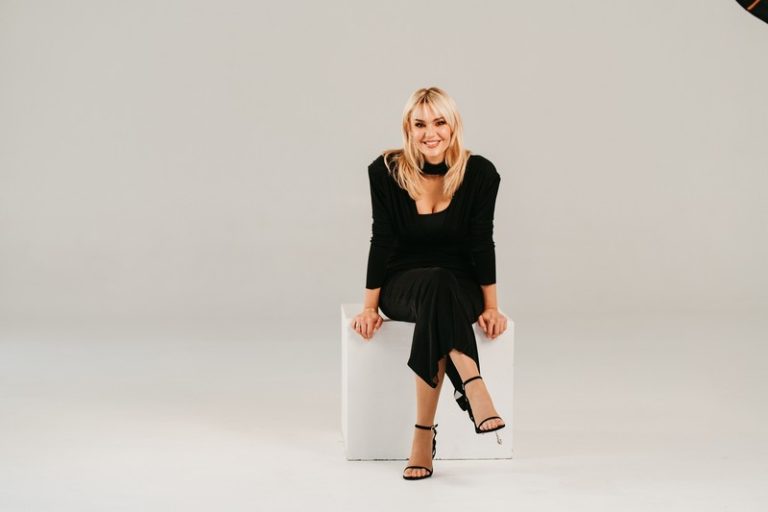 Lena Kovačević sedi na belom kubu u elegantnoj crnoj haljini, u studijskom ambijentu, nasmejana i okrenuta ka kameri.