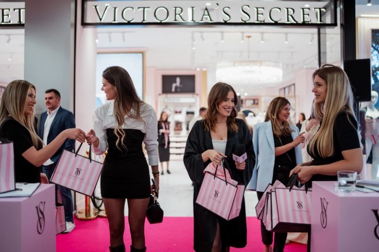 Kupci ispred prodavnice Victoria’s Secret sa roze kesama tokom svečanog otvaranja, uz crveni tepih i luksuzno uređen enterijer u pozadini.
