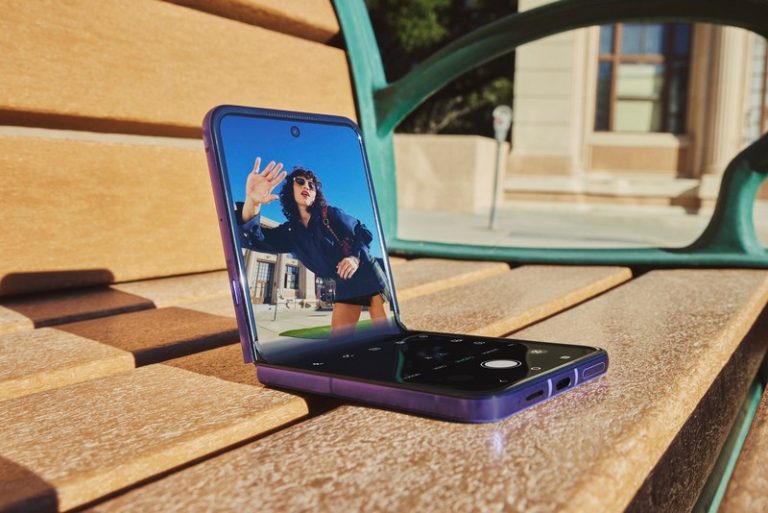 Preklopni telefon Motorola razr 70 ultra otvoren na klupi, sa prikazom kamere i osobe na ekranu u spoljašnjem okruženju.
