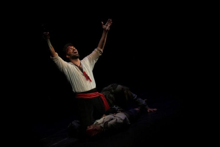 Glumac u emotivnoj sceni iz mjuzikla „Cigani lete u nebo“ sa podignutim rukama na tamnoj pozornici tokom izvođenja u okviru BELEF festivala u Beogradu.