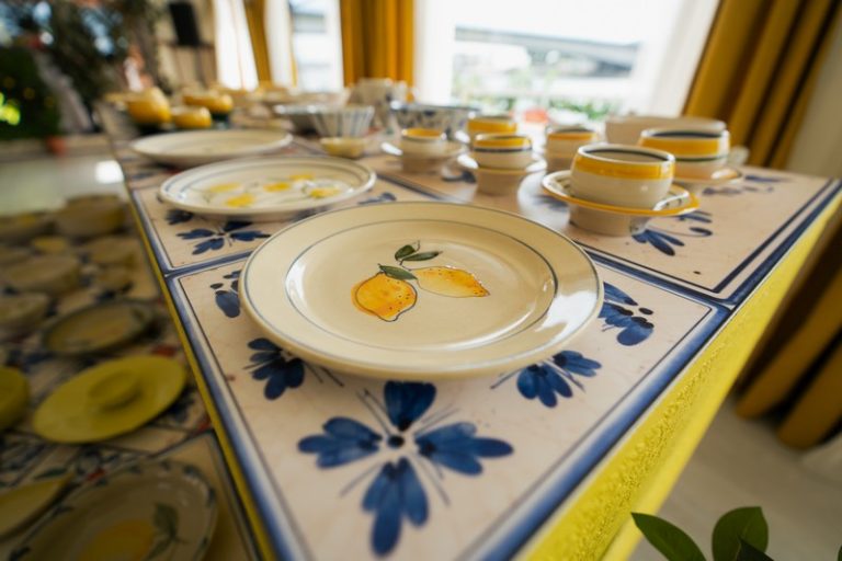 Ručно oslikani keramički tanjiri i šolje sa motivima limuna i plavih ornamenata postavljeni na dekorisan sto u mediteranskom, italijanskom stilu. Limoncellina